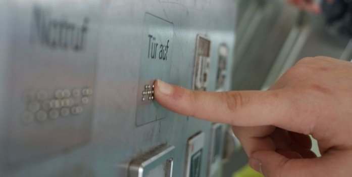 ein Finger drückt auf einen Aufzugknopf "Tür auf", der auch mit Braillezeichen beschriftet iste